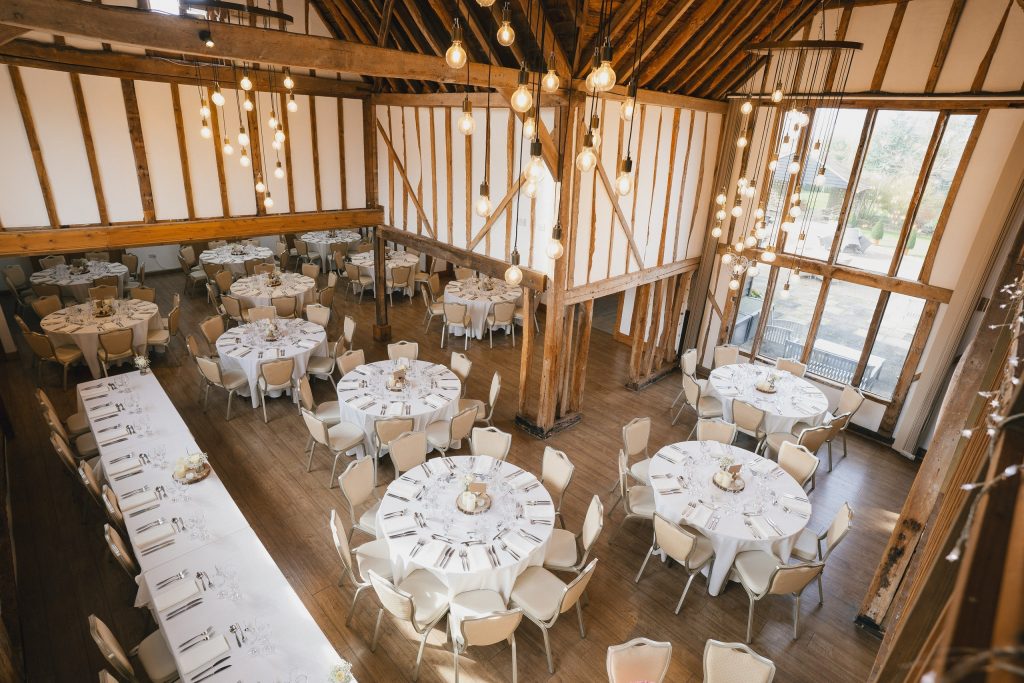 Wedding breakfast tables beneath exposed timber beams and hanging lights at Maidens Barn in Essex.